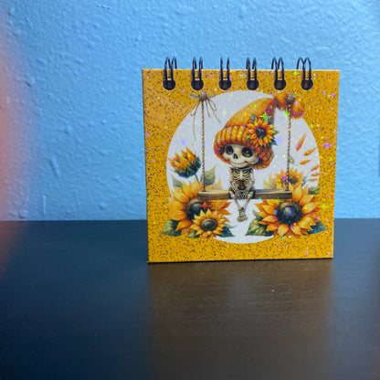 Small spiral-bound notebook with a skeleton in a sunflower hat design on a blue surface.