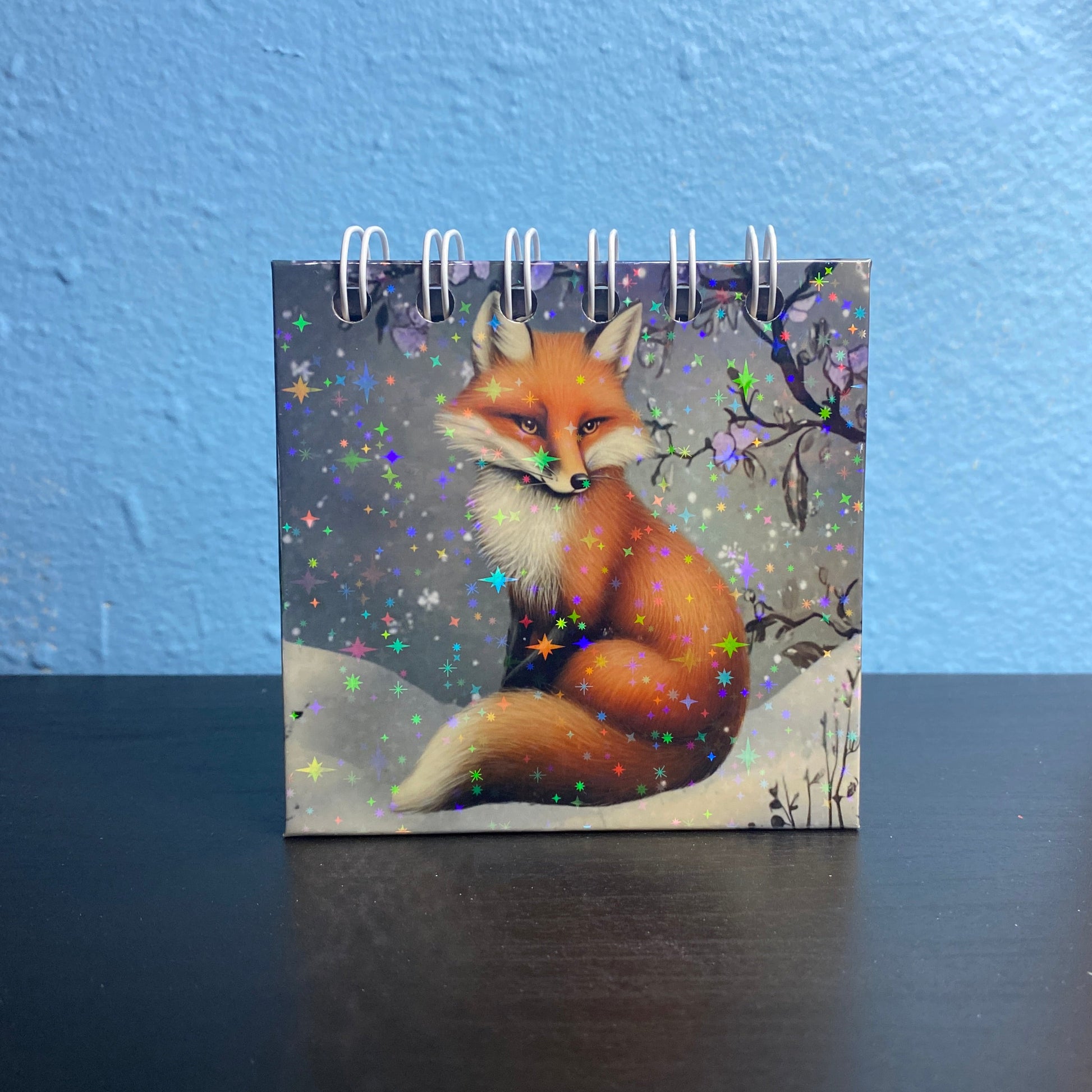 Small notebook with a fox design on a dark surface against a blue wall.