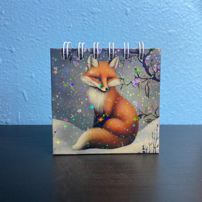 Small notebook with a fox design on a dark surface against a blue wall.