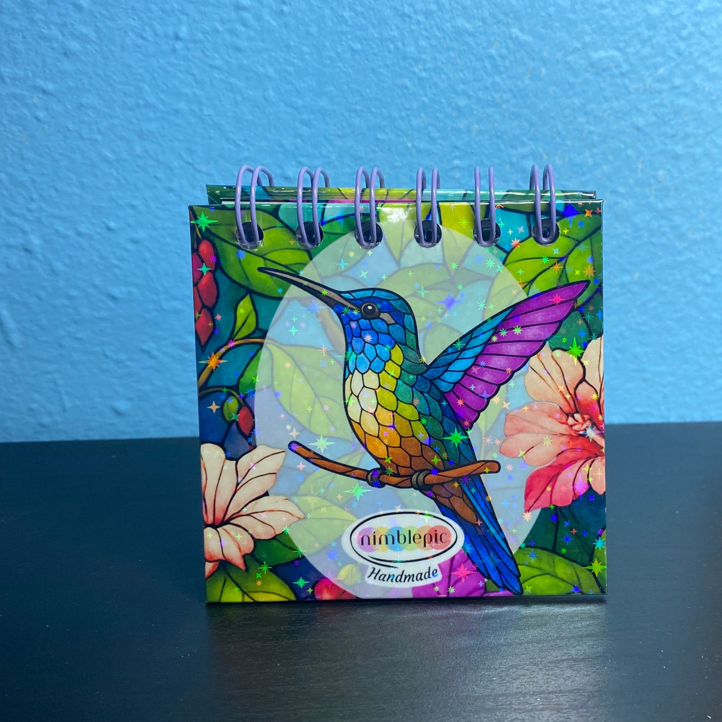 Colorful hummingbird-themed mini calendar on a dark surface with a blue wall background