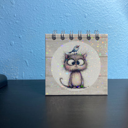 Small spiral-bound notebook with a cat illustration on a desk against a blue wall.
