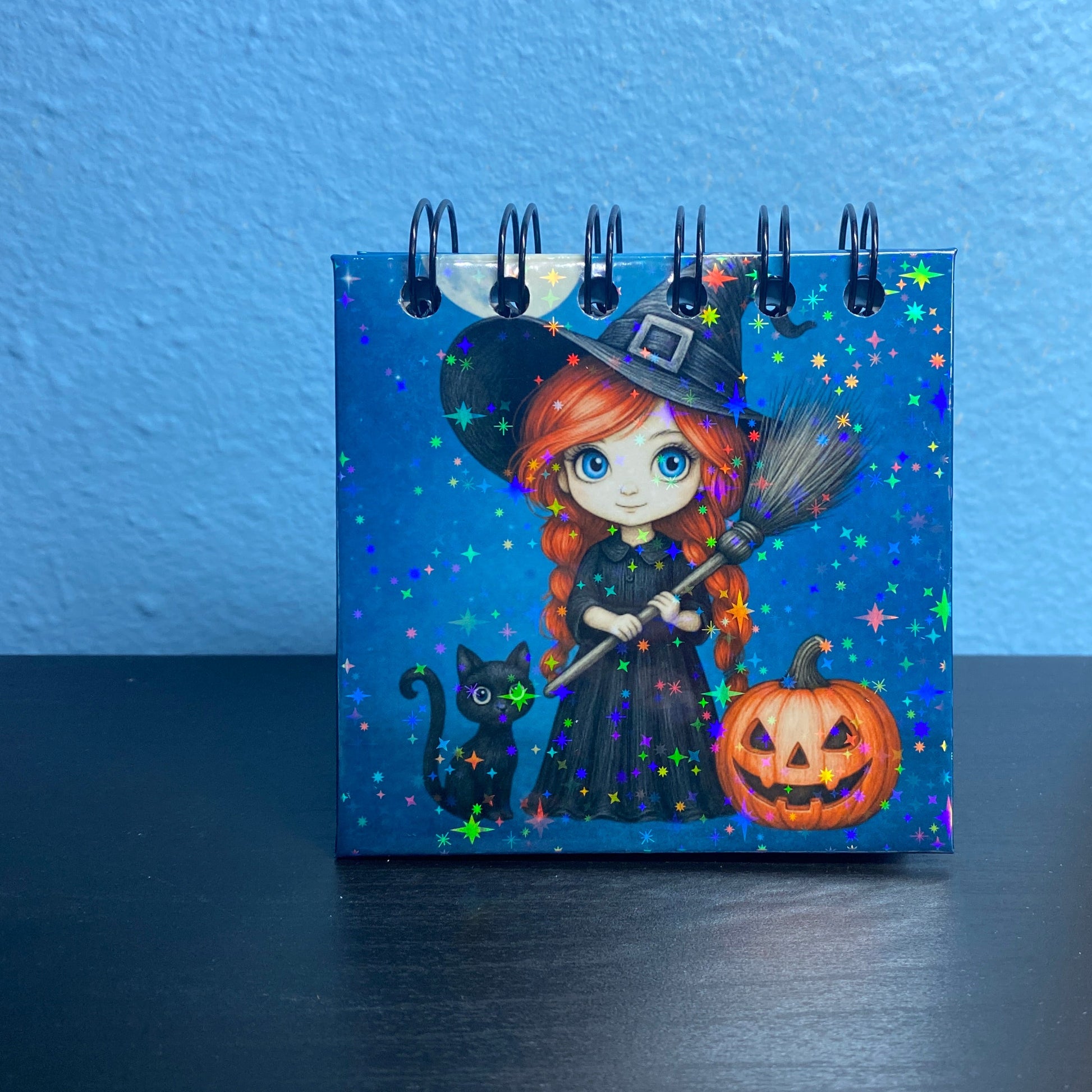 Small notebook with a Halloween-themed design featuring a witch, cat, and pumpkin on a blue background.