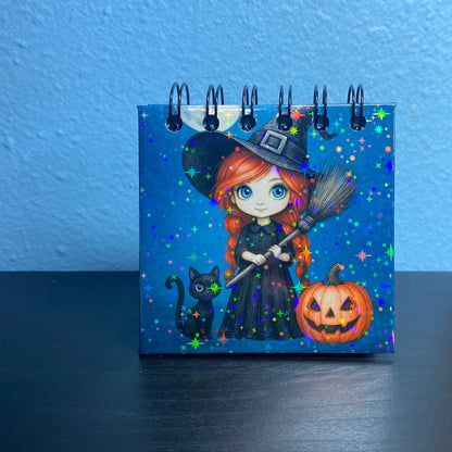 Small notebook with a Halloween-themed design featuring a witch, cat, and pumpkin on a blue background.